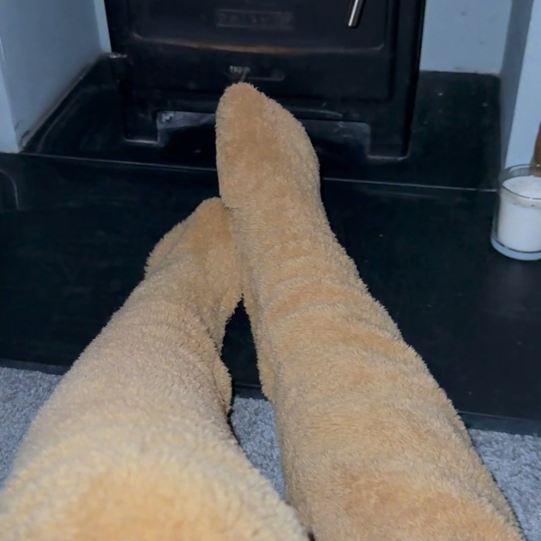 Crossed legs in camel teddy fleece over-the-knee leg warmers with covered feet, stretched towards a lit stove