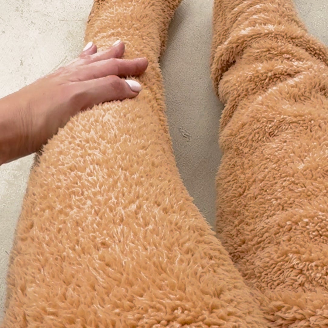 A hand smoothing the deep pile of a caramel fleece leg warmer worn over the knee, with the second leg alongside