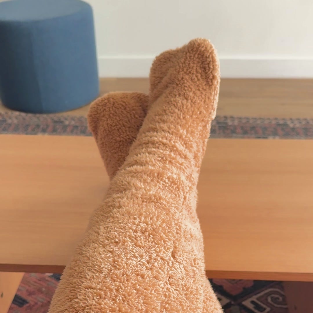 Both legs in caramel over-the-knee fleece leg warmers resting on a wooden bench above a patterned rug in a living room.