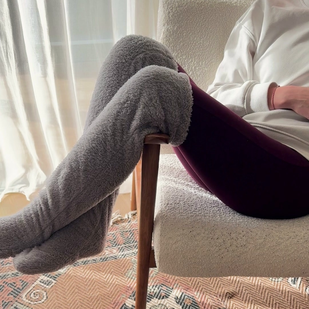 A woman sits in a cream armchair with grey plush over-the-knee leg warmers worn over maroon leggings, legs resting over the chair arm above a patterned rug.