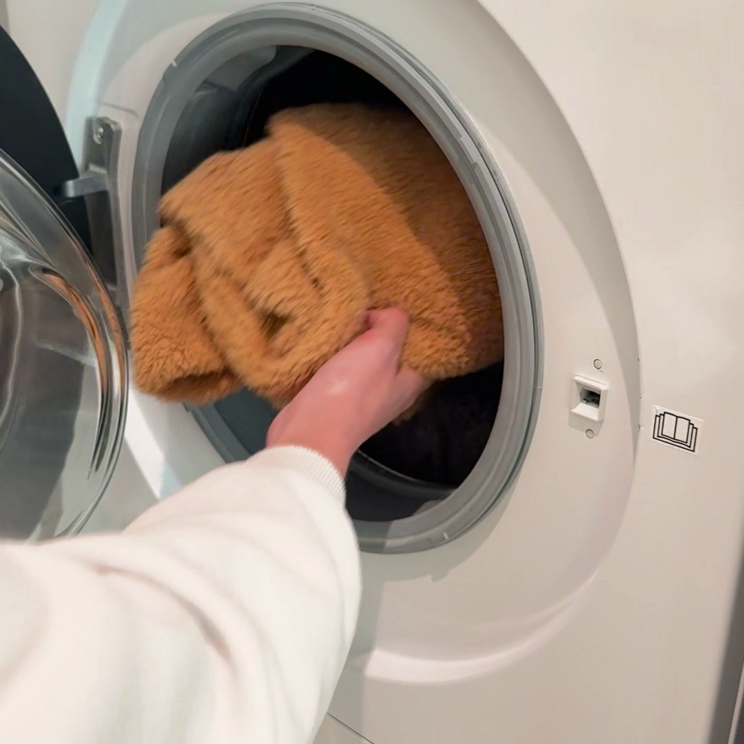 A hand pushing a pair of caramel plush fleece leg warmers into the drum of a white front-loading washing machine.